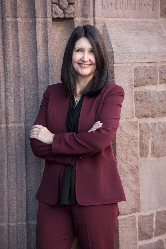 A woman with straight dark hair, wearing a burgundy suit and black blouse, stands smiling with arms crossed in front of a stone wall with carved details. She exudes confidence and professionalism.