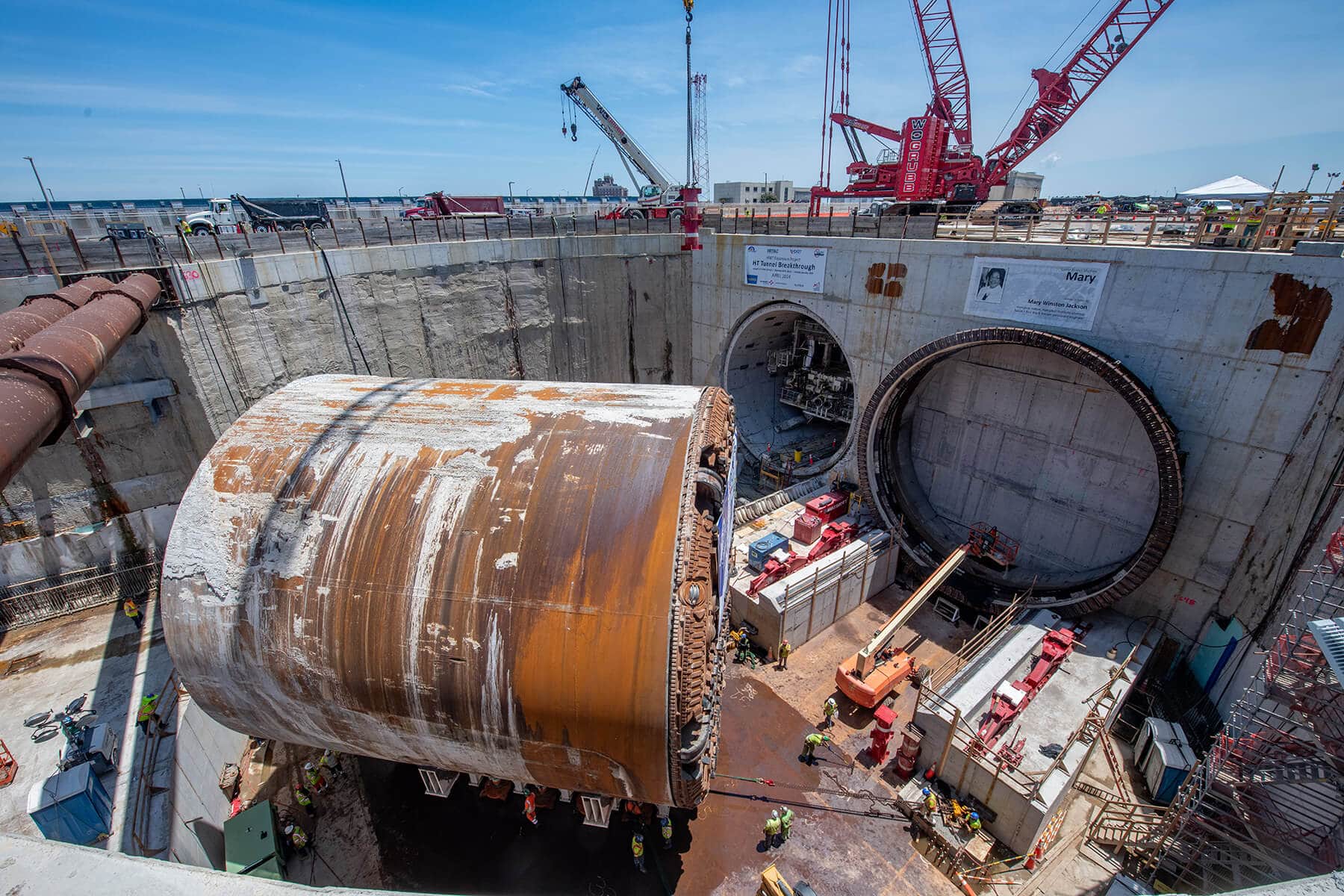 hampton-roads-tbm-turnaround A large tunnel boring machine is positioned at the edge of a circular concrete pit at a construction site. Workers in safety gear, cranes, and equipment surround the site. Two large tunnel entrances and various pipes are visible in the concrete wall.